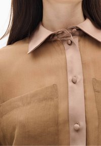 Chemise marron clair avec un col rose pâle, comportant deux boutons et deux poches avant. Texture douce, tissu légèrement transparent.