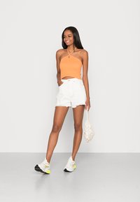 Top cropped laranja com nervuras e decote halter, calções de ganga brancos de cintura alta, e sapatilhas brancas com detalhes amarelos; segurando uma bolsa de rede bege.