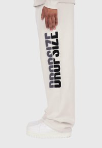 Gaiši bēši sporta bikses ar melnu tekstu "DROPSIZE" sānos. Izgatavotas no mīksta auduma, ar elastīgu jostu un ribotām aprocēm.