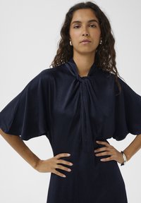 Temno modra obleka z ovratnikom na zavezovanje, kratkimi krilci v obliki netopirjih kril in teksturirano tkanino. Model stoji z rokami na bokih in nosi srebrne uhane.