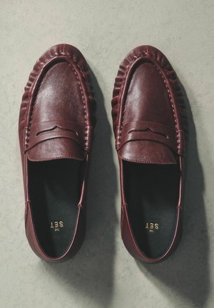 Burgundia nahast mokassiinid tekstuurse krae ja õmblustega detailidega, poolavatud disainiga, musta sisemusega ja kuldse brändinguga.