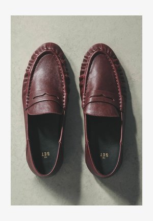 Viininpunaiset nahkaiset loafersit, joissa on teksturoitu kaulus ja ommellut yksityiskohdat. Kenkien muoto on puoliksi avoin, musta sisäpuoli ja kultaiset brändäystunnisteet.