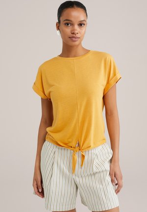 Camicia gialla a maniche corte con dettaglio anteriore annodato, abbinata a pantaloni corti bianchi plissettati con sottili strisce verdi. Tessuto liscio.