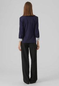 Blazer bleu marine avec une silhouette cintrée, manches trois-quarts et détail de manchette rayée, associé à un pantalon noir à jambes larges.