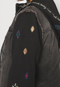 Svart denimjacka med färgglada broderade geometriska mönster på ärmarna och ett texturerat fransat kant. Dragkedja och kragdetaill synligt.