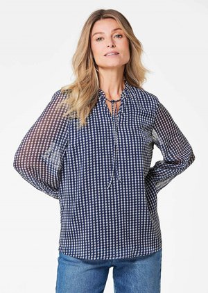 Marineblauwe blouse met geometrisch wit patroon, luchtige doorschijnende mouwen, striksluiting bij de hals, losse pasvorm en afgeronde onderrand.
