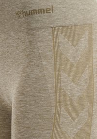 Beżowe legginsy sportowe mają teksturowane wykończenie, logo Hummel na talii oraz boczny pasek w wzór chevron.
