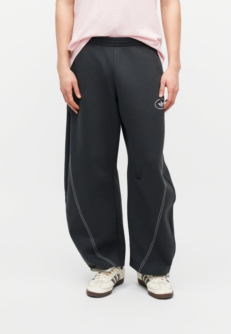 Άτομο που φορά μαύρο παντελόνι wide-leg με λευκές ραφές, λευκά αθλητικά παπούτσια Adidas και ανοιχτό ροζ πουκάμισο κομμένο στη μέση.