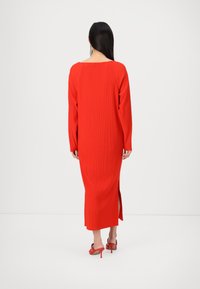 Femme aux longs cheveux foncés portant une robe midi rouge vif à manches longues côtelée et des sandales rouges à talons hauts, debout, de dos.