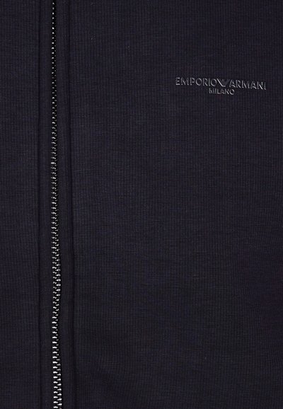 Cárdigan negro con cremallera de textura acanalada, que presenta una cremallera plateada y el logo en relieve "Emporio Armani Milano" en el lado izquierdo.