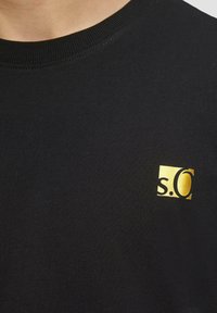 Schwarzes Baumwoll-T-Shirt mit Rundhalsausschnitt, mit einem kleinen goldenen Logo in der oberen linken Ecke. Glatte Textur, minimalistisches Design.