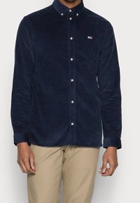 Tmavě modrá košile z manšestru s límečkem na knoflíky, s malým logem na hrudi, stříbrnými knoflíky a dlouhými rukávy s manžetami na knoflíky.