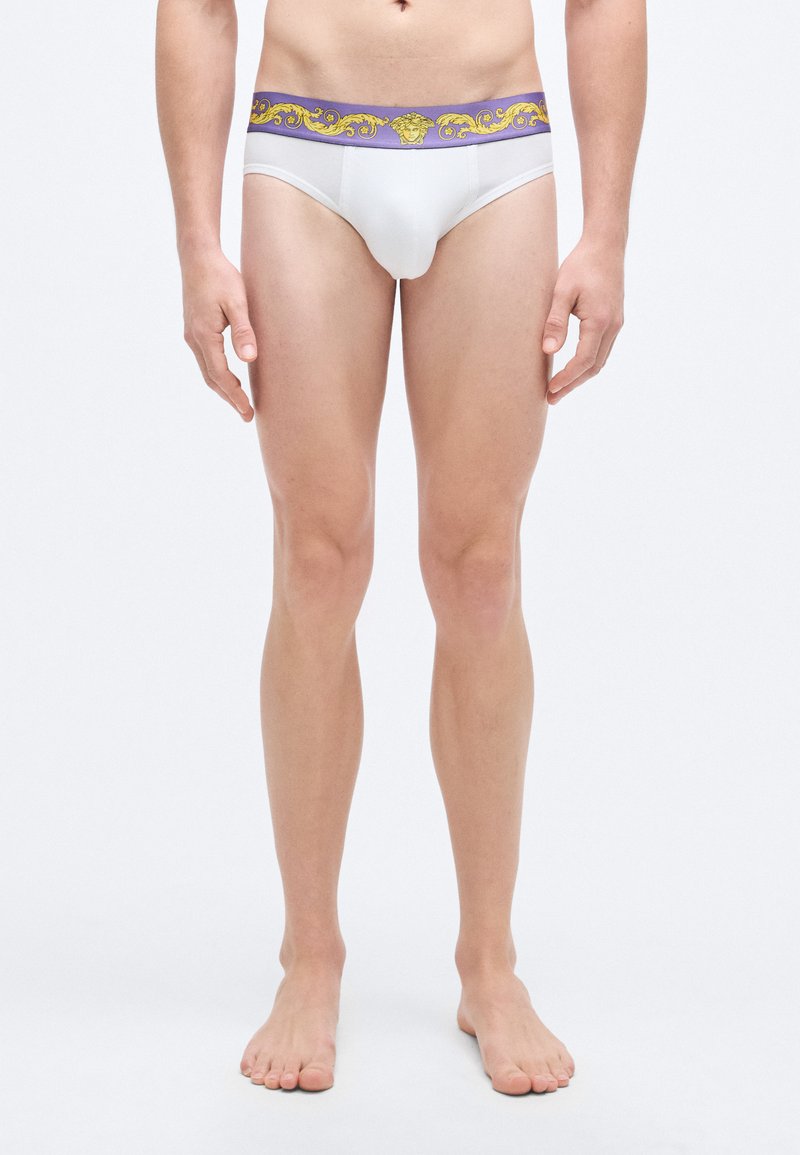 Λευκά σλιπ με μωβ ζώνη στη μέση που φέρει χρυσή διακοσμητική σχεδίαση. Το ύφασμα φαίνεται λείο και εφαρμοστό. Στυλ με ελάχιστη κάλυψη.