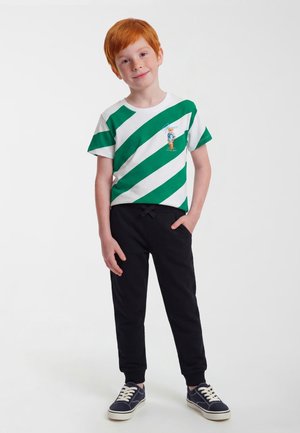 Red-haired boy standing with one hand in pocket, wearing white and green striped t-shirt, black pants, and navy sneakers.