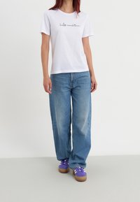 Vit bomullströja med svart kursiv text, tillsammans med ljusblå vida jeans och lila sneakers med vita snören och bruna sulor.