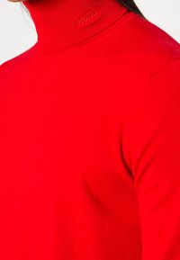 Pull rouge en maille à col roulé avec texture côtelée et logo détaillé sur le col. Vue en gros plan mettant en valeur le matériau et la coupe.