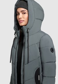 Gepolsterte graue Jacke mit hohem Kragen, verstellbarer Kapuze, Reißverschluss- und Druckknopfverschlüssen sowie schwarzen Akzentdetails, darunter ein Logo-Patch und eine Seitentasche.