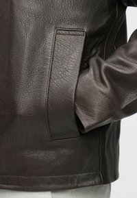 Tuvplāns ar tumši brūnas ādas jaka kabatu, kurā daļēji ielikta roka, redzama teksturēta āda un šūtas šuves.