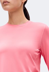 Κοντινό πλάνο μιας osoby wearing a smooth, μακρυμάνικη ροζ μπλούζα με στρογγυλή λαιμόκοψη, με σκούρα μαλλιά δεμένα σε αλογοουρά.