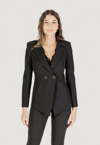 Černý dvouřadý blejzr s špičatými klopami, zlatými kovovými detaily a přiléhavým střihem, kombinovaný s černým topem a slim fit kalhotami.