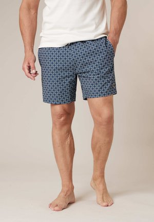 Homme portant un short bleu marine à motifs et un t-shirt blanc, debout pieds nus avec une main dans la poche, sur un fond neutre.
