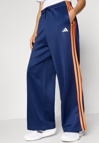 Marineblå vidbente joggebukser med oransje sidestriper, elastisk midje med snøring og hvit logo på venstre lår.