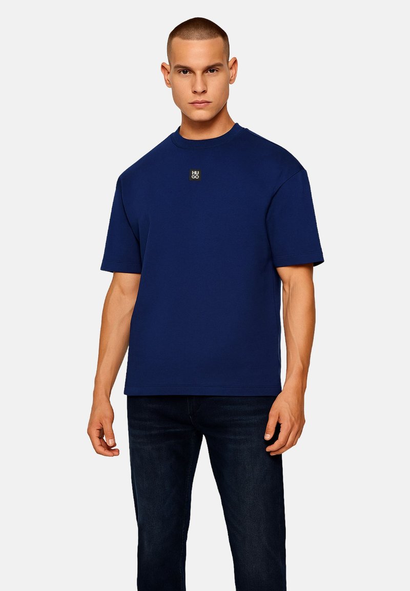 Camiseta de manga corta azul marino en tejido suave con cuello redondo. Presenta un pequeño logo dorado en el pecho y un corte holgado.