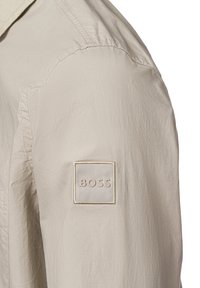 Beige lichtgewicht jasje met een gestructureerde stof, voorzien van een schouderpatch met de opdruk "BOSS" in reliëfletters en een klassiek kraagontwerp.