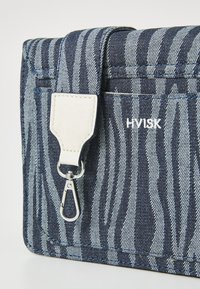 Bolso mensajero de mezclilla con rayas de cebra en azul y gris, que presenta un acento de correa de cuero blanca y herrajes plateados. Logo: "HVISK."