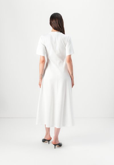 Weißes, knielanges Kleid mit kurzen Ärmeln, glatter Textur, einfachem Design und geradem Schnitt. Getragen mit schwarzen, offenen High Heels.