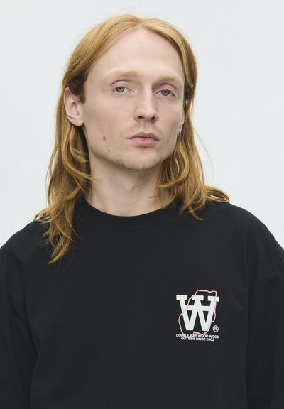 Jeune personne aux longs cheveux roux portant un sweat-shirt noir avec un logo graphique blanc et rose sur la poitrine sur fond uni.