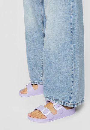 Fioletowe gumowe sandały z dwoma regulowanymi paskami i klamrami, zestawione z jasnoniebieskimi dżinsami o luźnym kroju i prostych nogawkach.