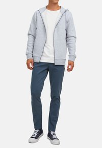 Light gray zip-up hoodie with a drawstring hood, worn over a white t-shirt. Paired with blue pants and navy sneakers featuring white soles.