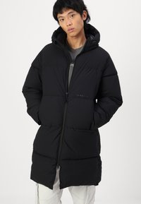 Abrigo acolchado negro con capucha, con cierre de cremallera, diseño acolchado y bolsillos laterales. Textura suave, silueta hasta la rodilla.