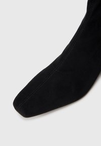 Bota ankle preta em camurça com bico apontado, apresentando detalhes de costura subtil e uma textura suave, angulada para uma vista lateral.