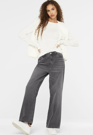 Junge Frau mit lockigem Haar trägt ein weißes Langarmshirt, weiße ärmellose Weste, lockere dunkelgraue Jeans und weiße Sneakers, posiert vor weißem Hintergrund.