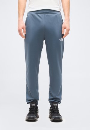 Mees, kes kannab siniseid North Face dressipükse ja musti North Face matkajalatseid, seisab käed rahulikult külgedel.