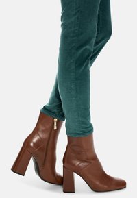 Brown leather ankle boots with a square toe, chunky heel, and side zipper detail, paired with green velvet skinny pants.