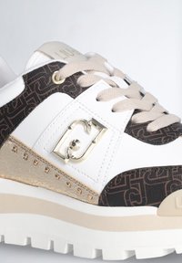 Zapatilla blanca con lateral de patrón marrón, acentos beig, herrajes dorados y panel metálico texturizado. Suela gruesa con un dibujo pronunciado.