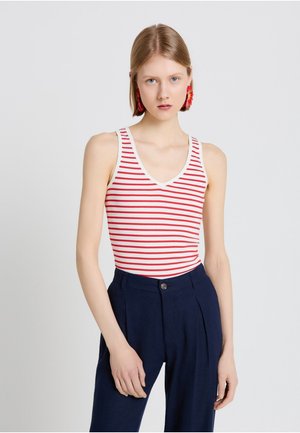 Giovane donna con capelli biondi corti che indossa una canottiera a righe rosse e bianche, pantaloni blu scuro a vita alta e orecchini a forma di fiore rossi, in piedi davanti a uno sfondo bianco.