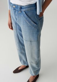 Light blue patchwork denim jeans worn with hands in pockets and brown flats, paired with a white top and light blue cardigan.