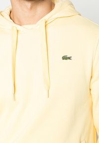 Man bär en blekgul huvtröja med dragsnören och en liten grön krokodil-logotyp på bröstet.