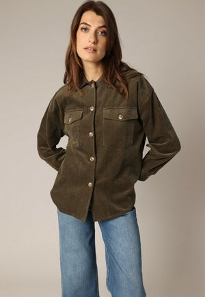 Chemise en velours côtelé vert olive avec deux poches poitrine, présentant un tissu texturé et un col classique. Portée avec un jean bleu clair.