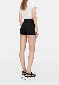 Pantalones cortos negros de talle alto con bolsillos traseros, combinados con una blusa de punto blanca y sandalias negras con suelas gruesas blancas.
