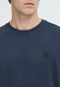 Camiseta de algodón azul marino con cuello redondo y un logotipo geométrico negro bordado en el lado izquierdo del pecho. Textura suave, diseño sencillo.
