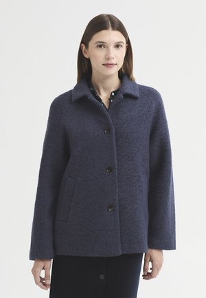 Femme portant un manteau en laine bleu marine avec trois boutons, debout devant un fond uni clair.