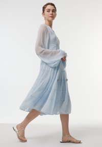 Robe bleu clair à étages avec manches longues transparentes et corsage ajusté, avec un ourlet fluide. Des sandales argentées à brides complètent la tenue.