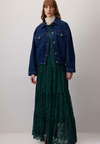 Giacca di jeans con due tasche sul petto, indossata sopra un lungo vestito verde di pizzo con strati a balze e bordi ondulati. Sciarpa leggera attorno al collo.