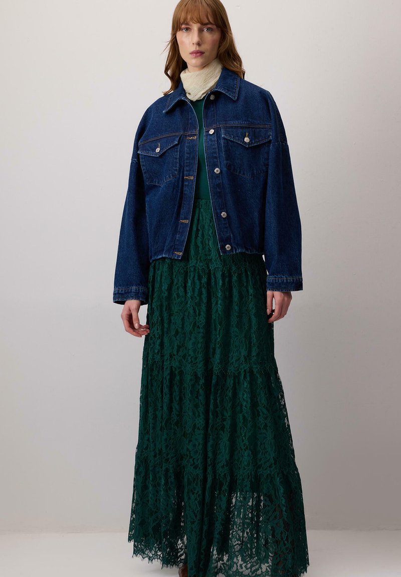 Giacca di jeans con due tasche sul petto, indossata sopra un lungo vestito verde di pizzo con strati a balze e bordi ondulati. Sciarpa leggera attorno al collo.