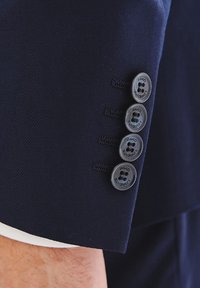 Rękaw marynarki w granatowym kolorze z czterema ciemnymi guzikami ozdobionymi nazwą marki, widocznymi szczegółami przeszyć oraz kontrastowym białym wykończeniem mankietów.